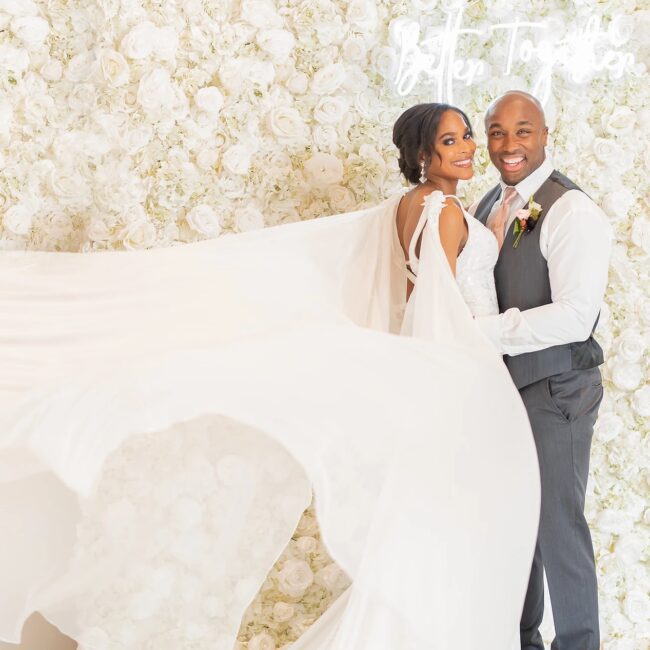 Bride and groom posing in front of a white floral wall, her long gown train flowing