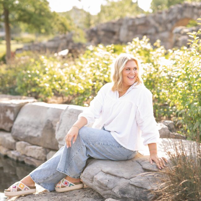 Senior girl in a white blouse sitting on a stone wall in a garden