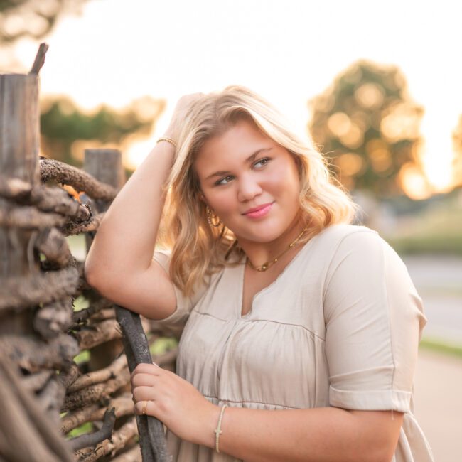 Senior girl leaning against a rustic wooden fence at golden hour