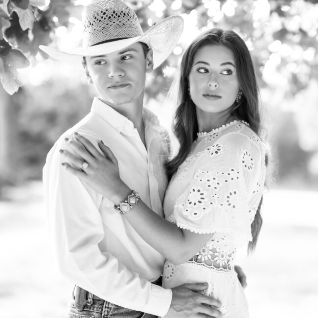 Black and white portrait of a couple standing close under the trees, him in a cowboy hat
