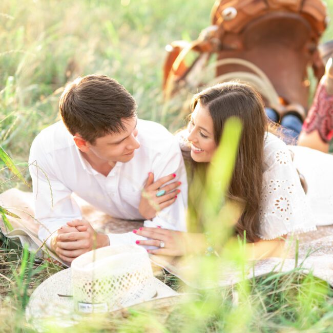 Couple lying in tall grass smiling at each other with a hat and boots nearby