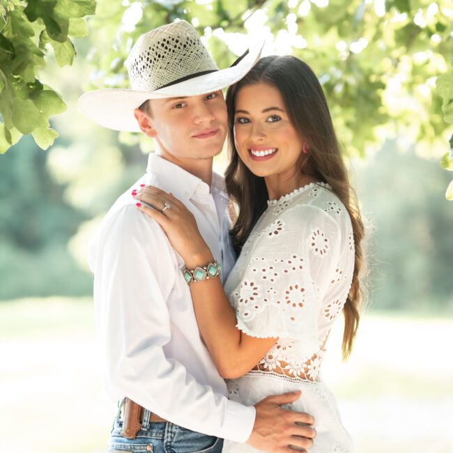 Engaged couple embracing, him in a cowboy hat, her in a white eyelet dress